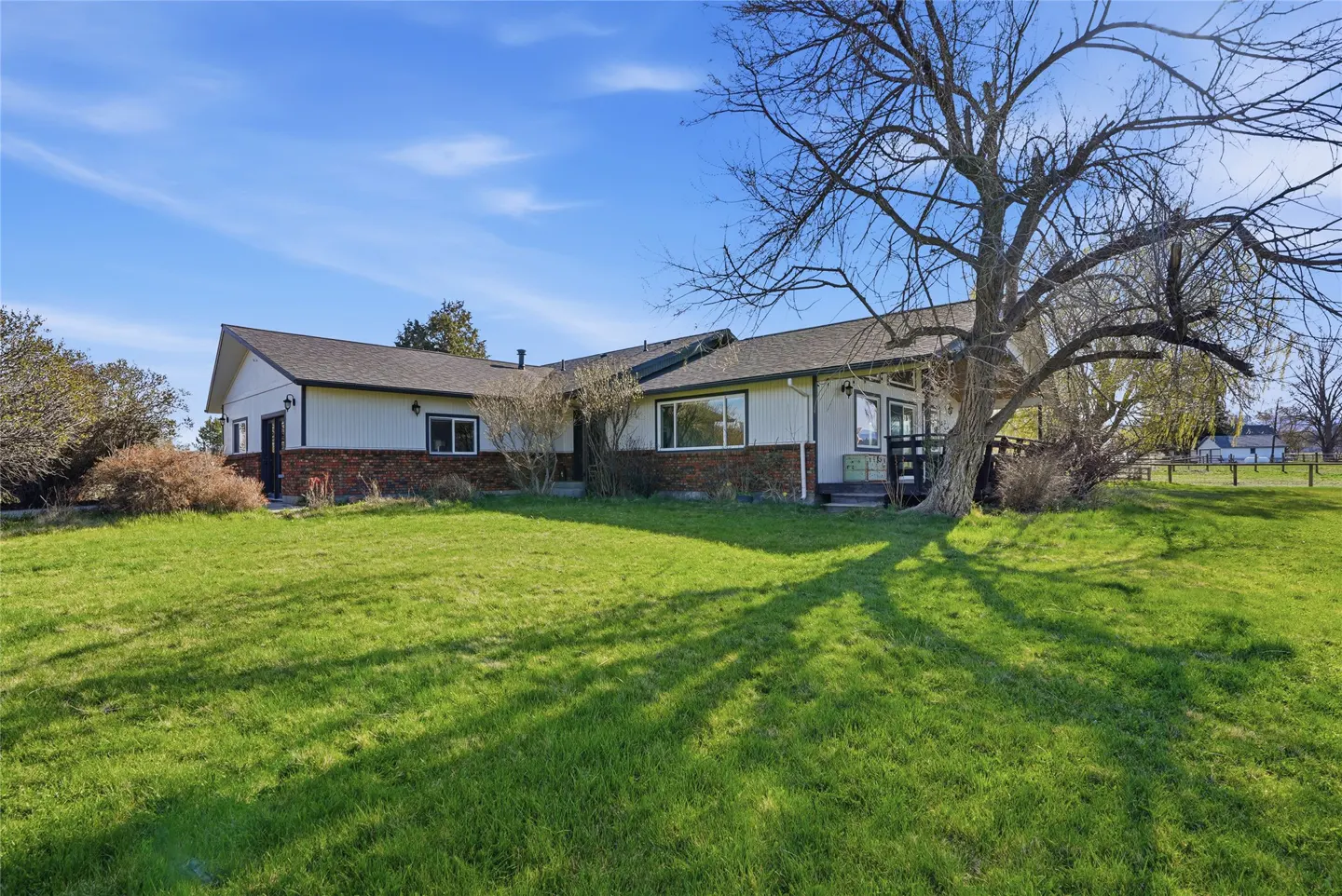 A ranch-style house with white siding and a brick foundation sits on a large green lawn under a blue sky. A large tree casts shadows on the grass.