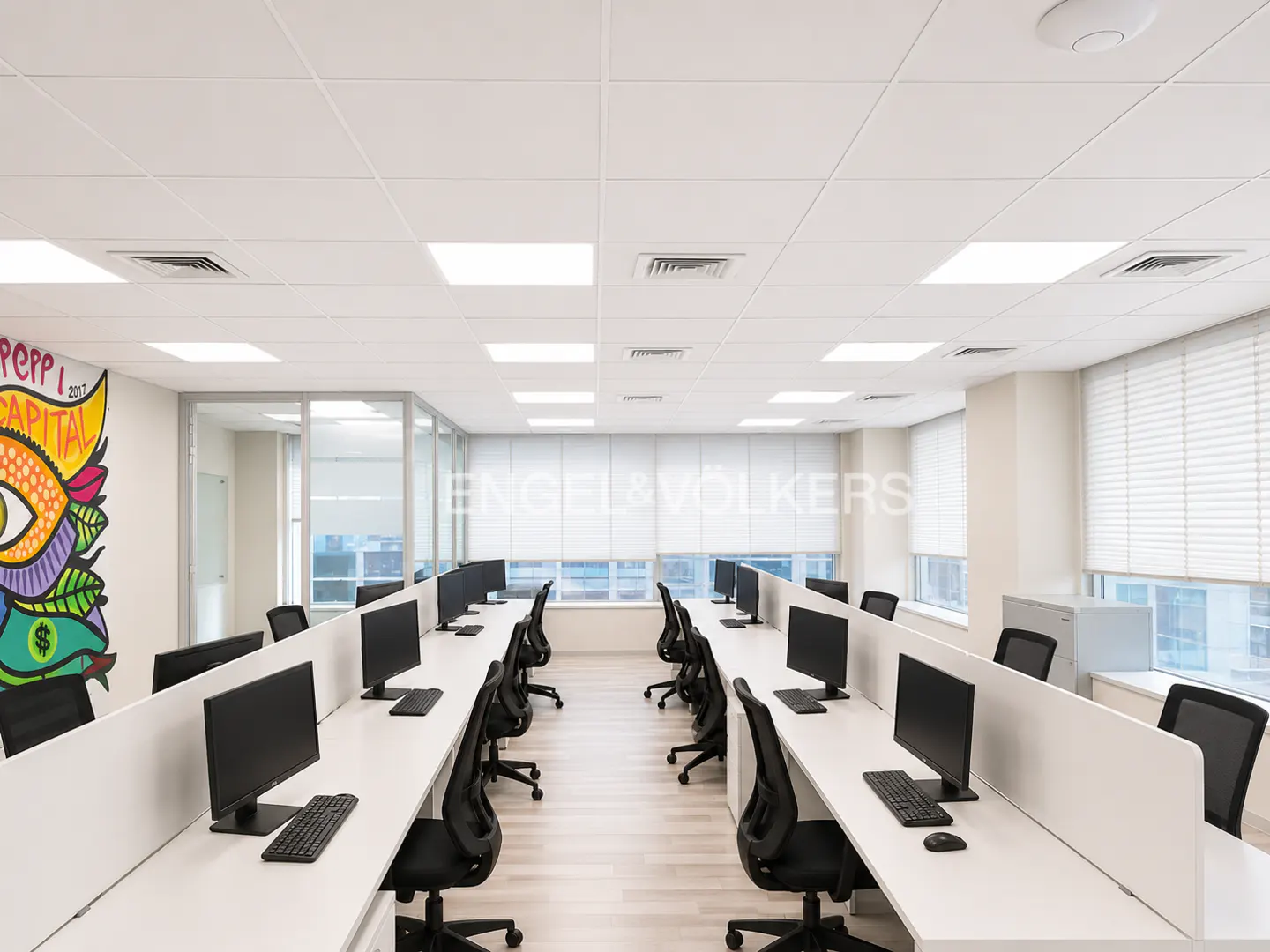 Bright office space with rows of white desks, black chairs, and computers. A colorful mural is on the left wall.