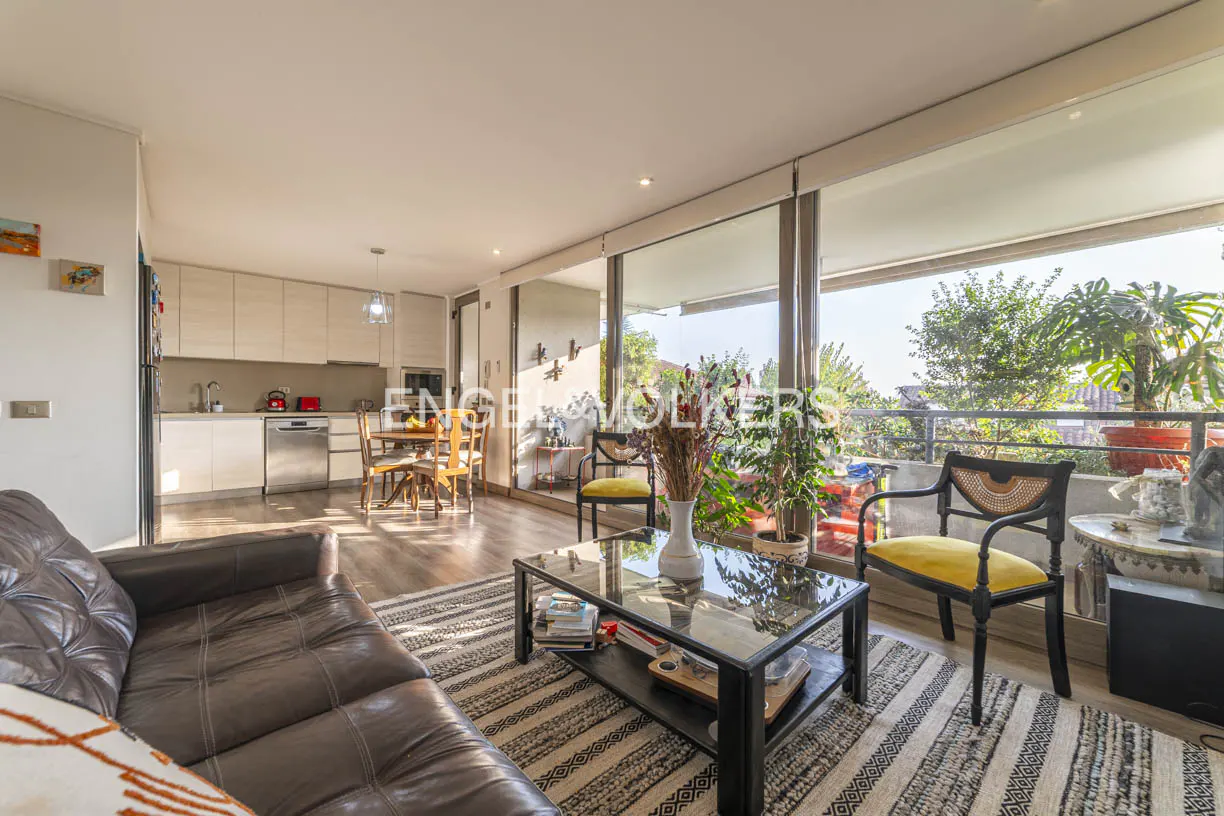 Bright living room with brown leather sofa, striped rug, glass coffee table, and balcony with plants. Kitchen in background.
