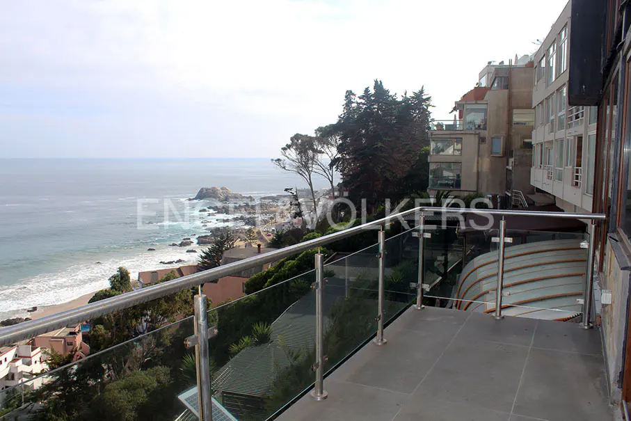 Balcony view of the ocean with rocks and beach below. Stainless steel and glass railing. Buildings and trees in the background.