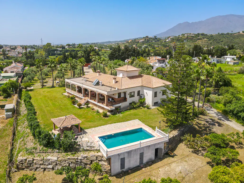 Aerial view of a large, white house with a terracotta roof, a pool, and lush green landscaping. A mountain is visible in the background.