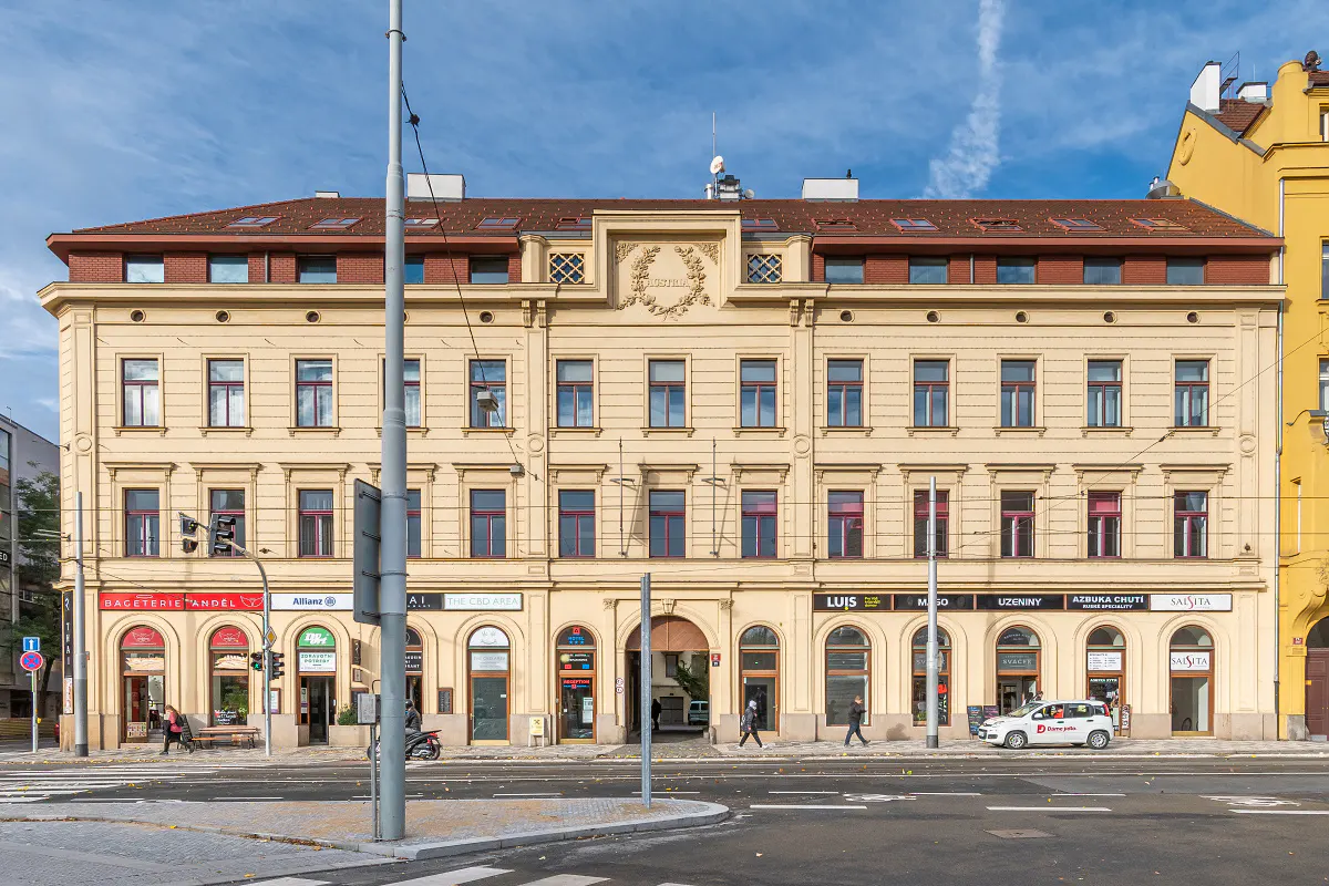 A four-story, light yellow building with red window frames and a red tile roof, street-level shops, and a blue sky.