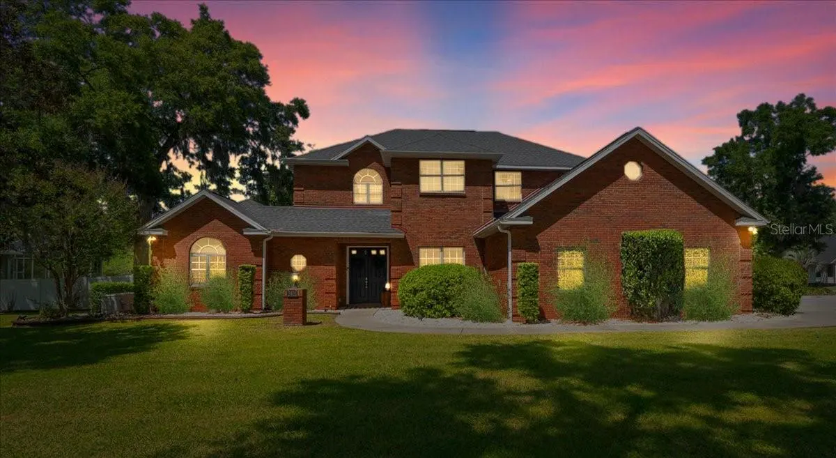 Two-story red brick house with a dark roof, green lawn, and pink and blue sunset sky.