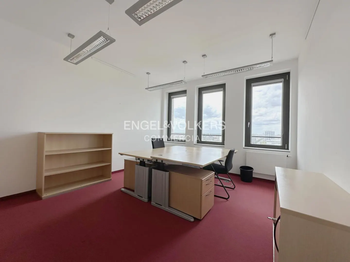 Bright office space with red carpet, white walls, and three windows. A large desk, chairs, and a wooden bookshelf furnish the room.