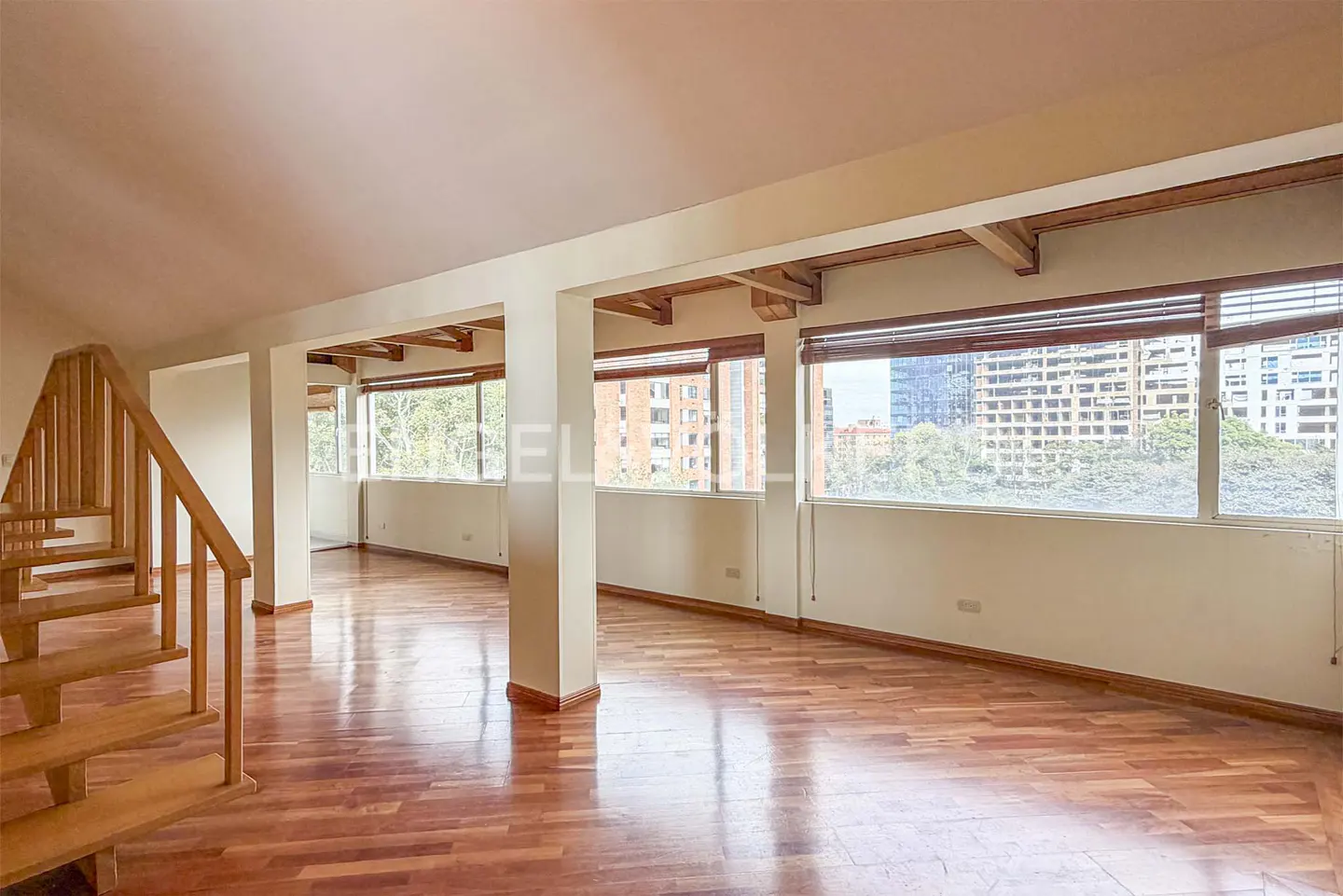 Bright, open living space with wood floors, white walls, and large windows with city views. Wooden staircase on the left.