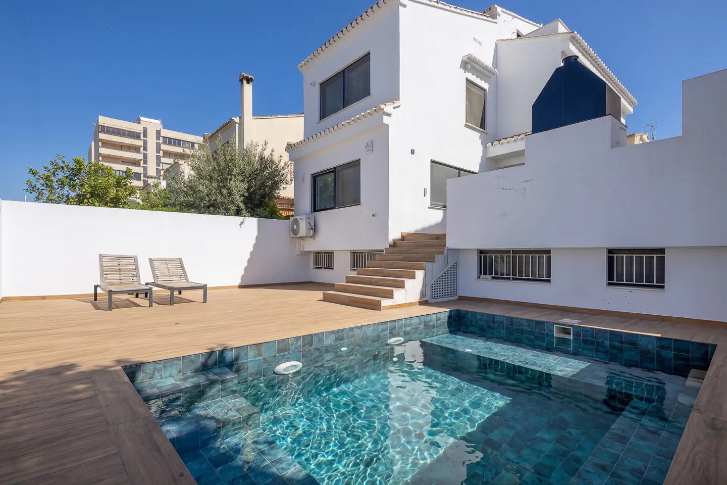 Backyard view of a two-story white house with a blue-tiled pool and wooden deck. Two lounge chairs sit on the deck. Blue sky.