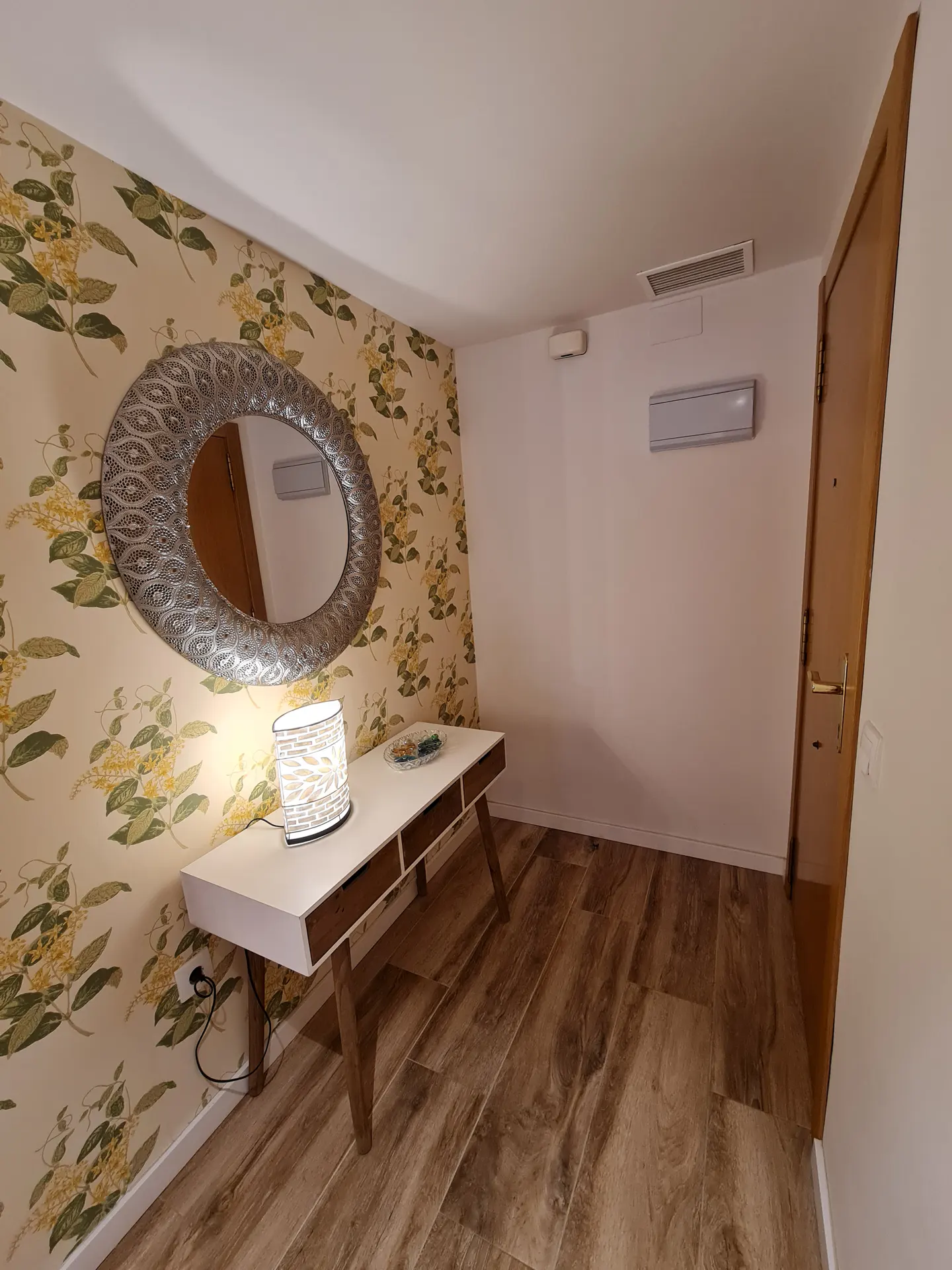 Entryway with wood-look tile floor, floral wallpaper, white console table with lamp, and decorative silver mirror.