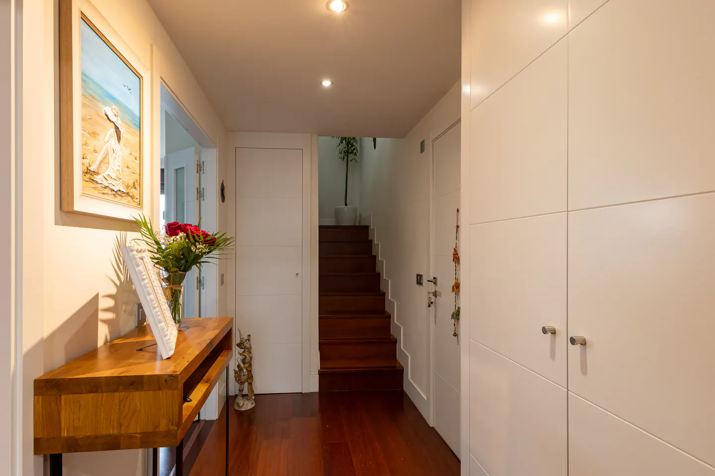 Hallway with wood floors, white walls, and recessed lighting. A wood table with flowers and art is on the left. Stairs lead up in the center.