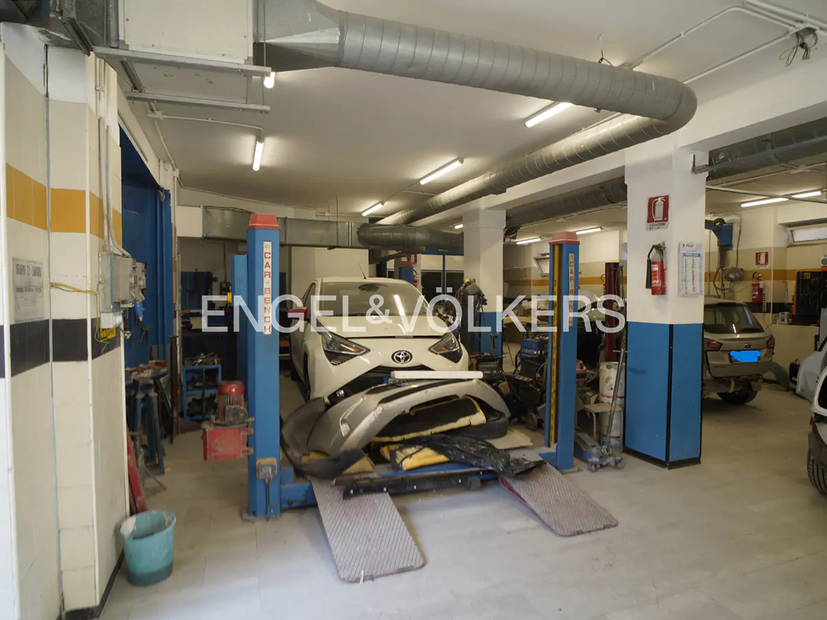 Auto repair shop interior with a white car on a blue lift, tools, and gray ductwork. Engel & Völkers logo visible.