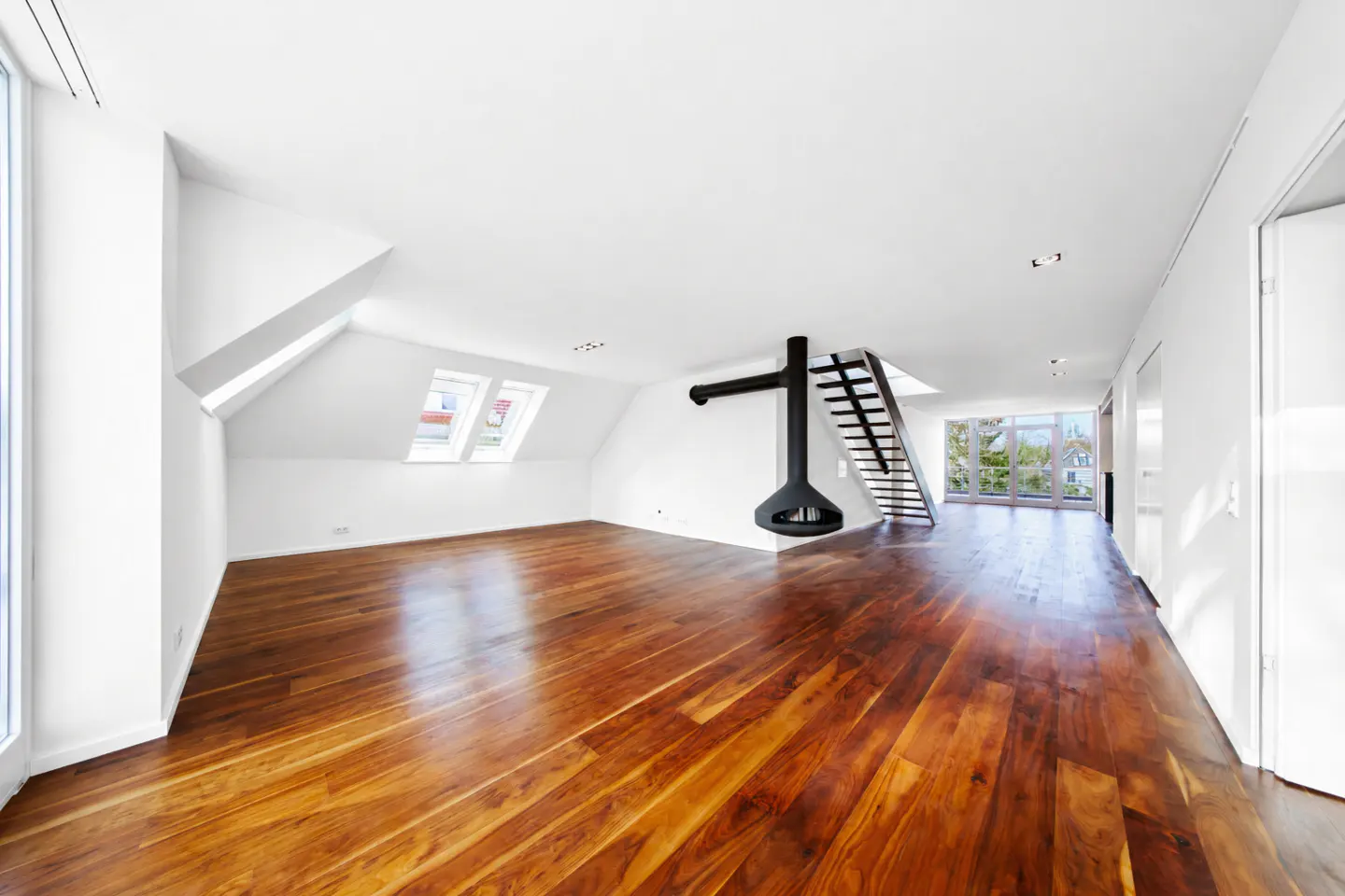 Bright, empty room with wood floors, white walls, and a black hanging fireplace next to a staircase. Skylights and a large window provide natural light.