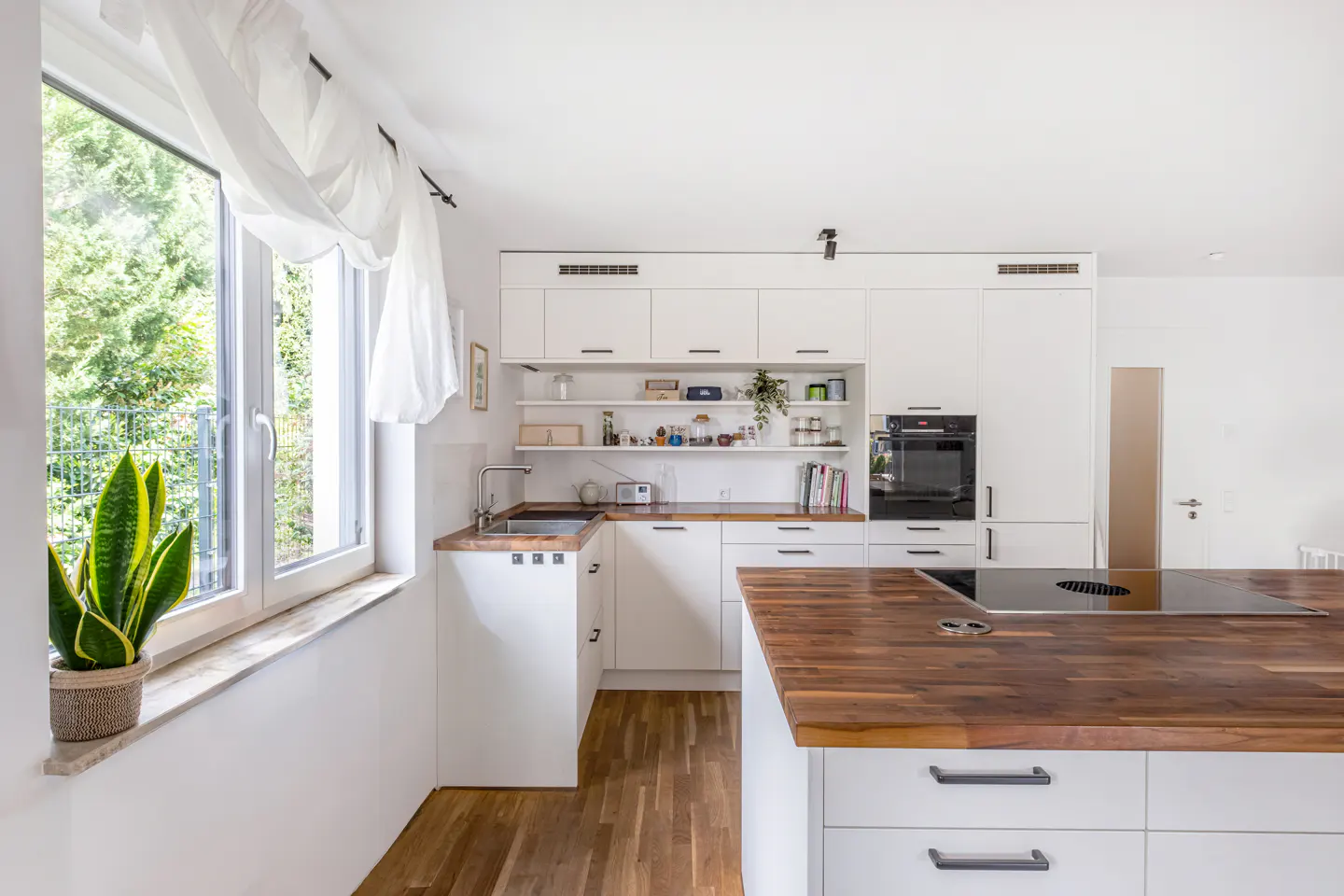 Modern white kitchen with wooden countertops, a central island, and a window with a snake plant on the sill.