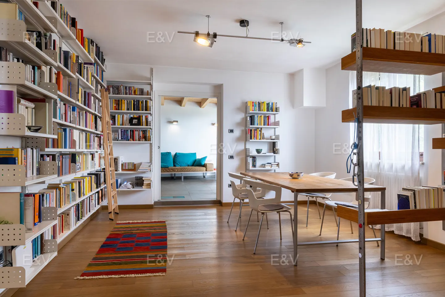 Bright home library with floor-to-ceiling bookshelves, a wooden dining table, and a colorful rug on hardwood floors.