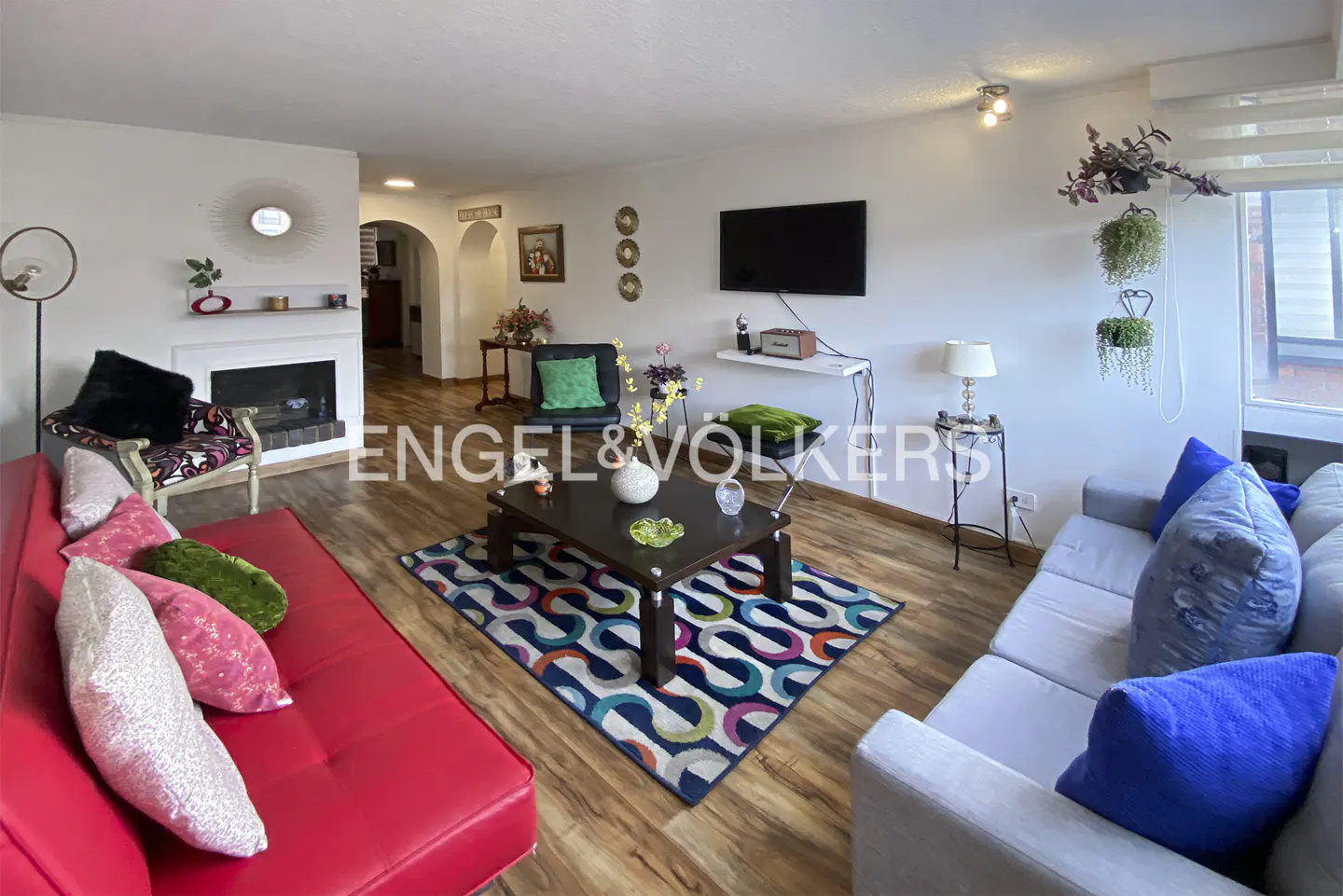 Bright living room with a red sofa, grey couch, colorful patterned rug, fireplace, and wall-mounted TV on wood floors.