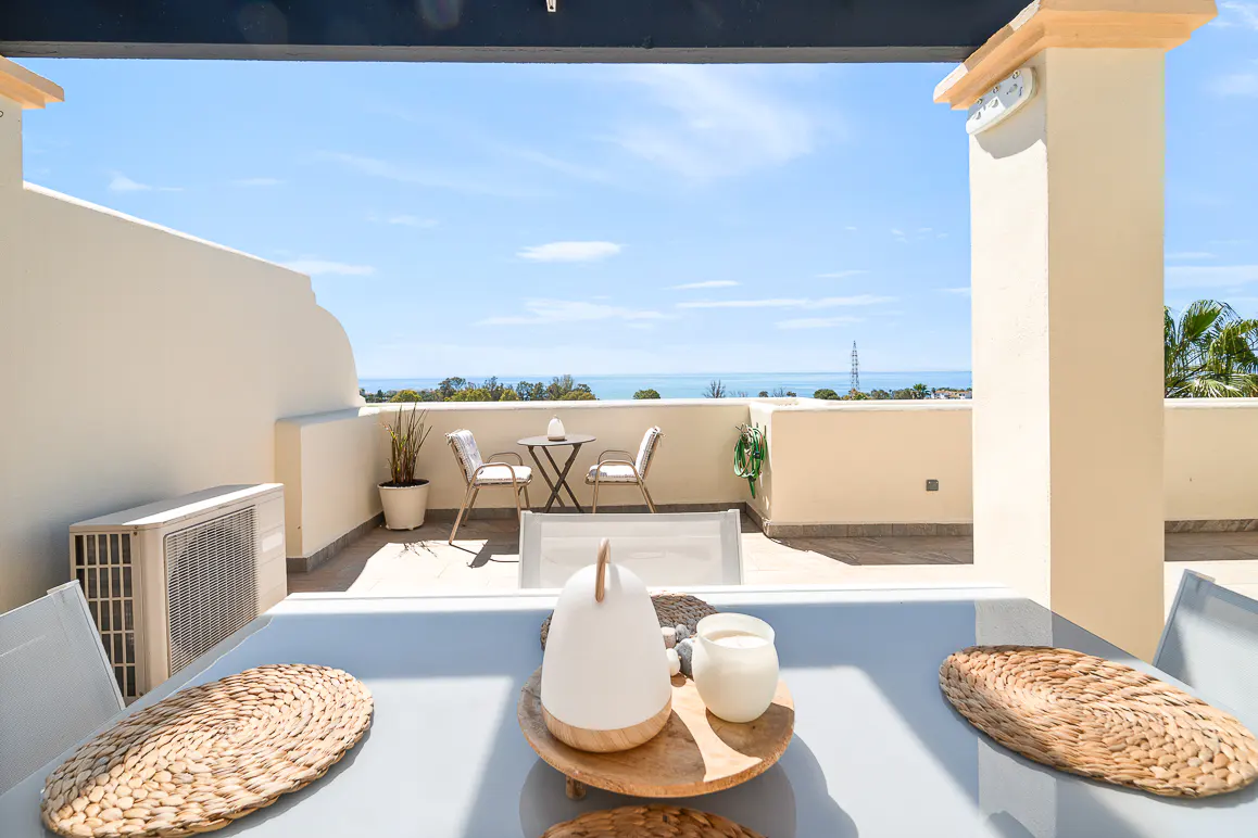 Balcony view with table setting, placemats, and decor. Ocean and blue sky in background. Cream walls and outdoor furniture.