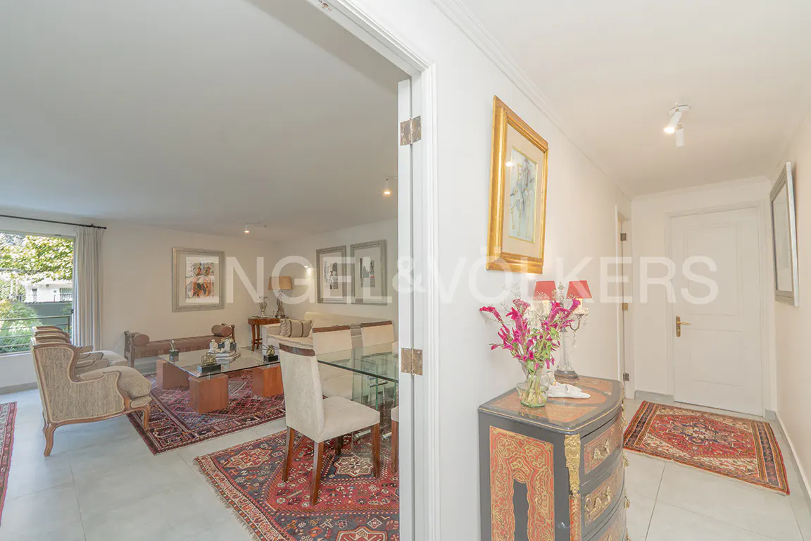 A bright, open-concept living and dining area is viewed through a doorway. A gold-framed painting hangs above a decorative chest of drawers.