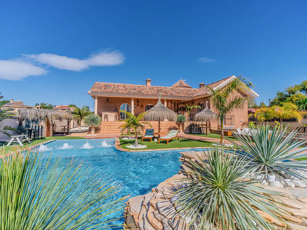 A sunny view of a luxury home with a pool, thatched umbrellas, and palm trees. The house is brick with a red tile roof.