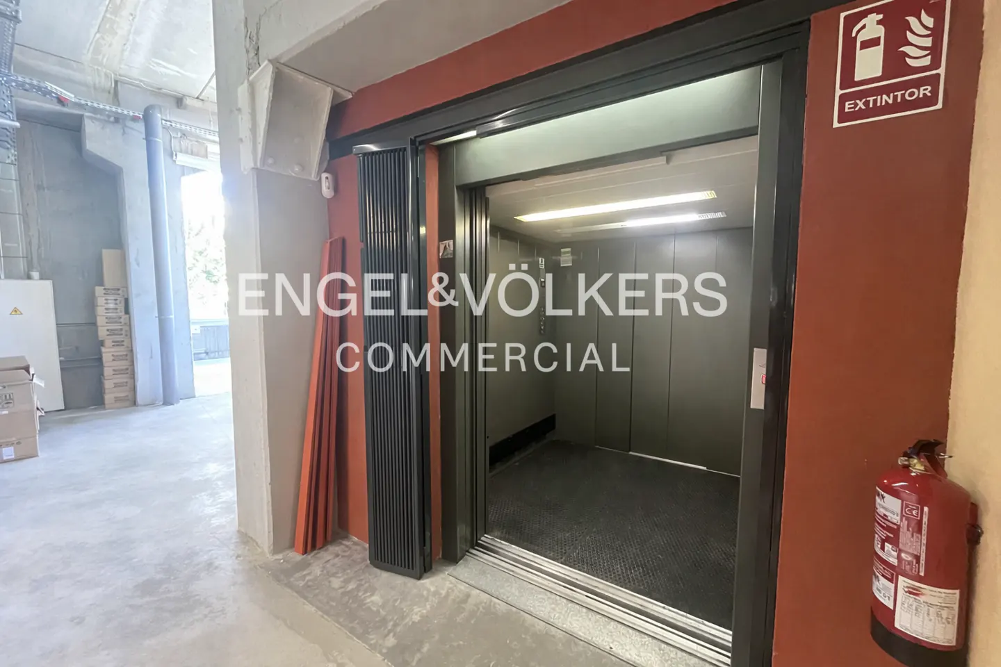 A commercial elevator with an open door, showing the interior, next to a red wall with a fire extinguisher.