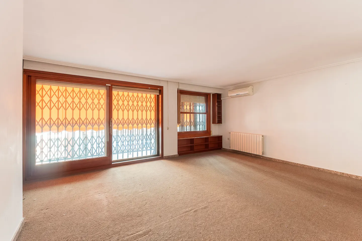 Empty room with brown carpet, white walls, and wood-framed windows with metal security grilles. A radiator and AC unit are on the right wall.