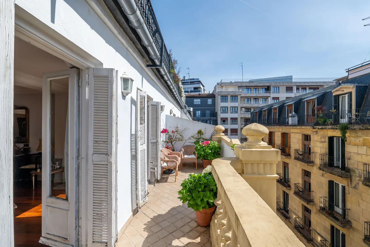 Balcony with potted plants and wicker chairs, overlooking city buildings under a blue sky. White shutters are partially open.
