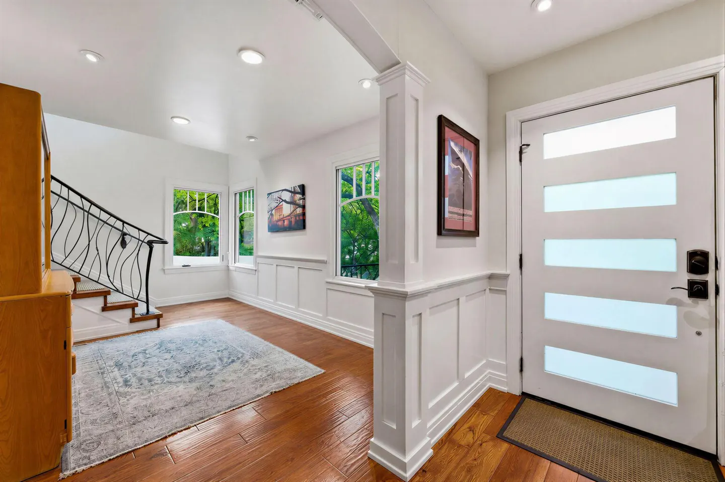 Bright entryway with hardwood floors, white walls, and a modern white door with glass panels. A staircase and rug are visible.