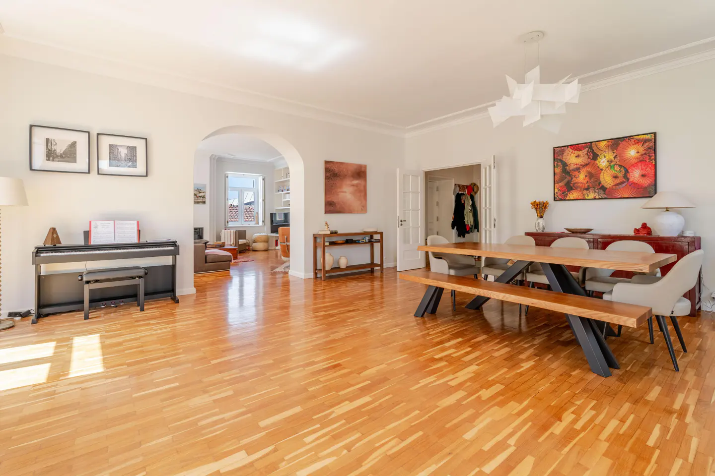 Bright, open-concept living and dining room with hardwood floors. A piano sits near framed art, while a wooden table and bench anchor the dining area.