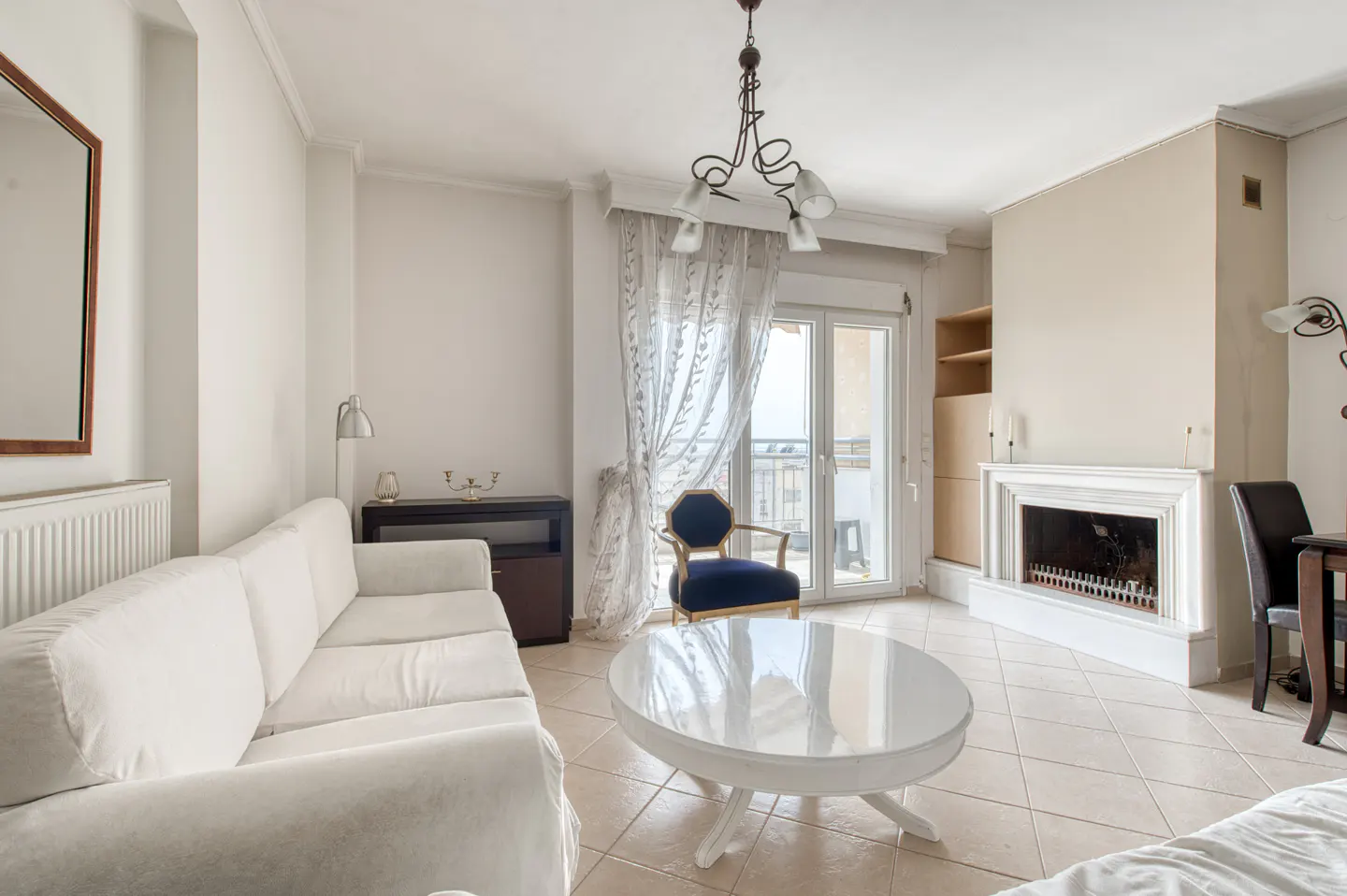 Bright living room with white sofa, oval table, blue chair, and fireplace. Balcony doors with sheer curtains let in natural light.