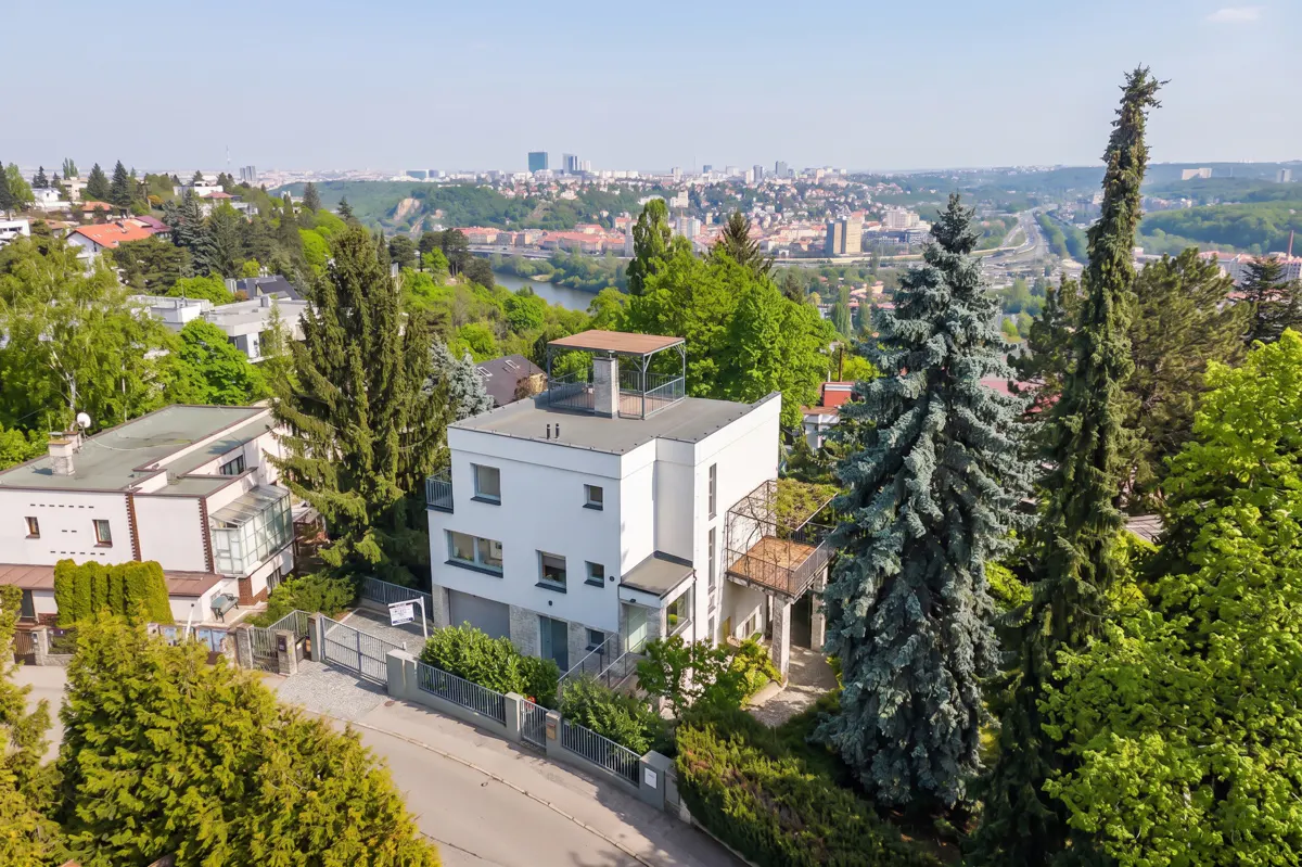 Aerial view of a modern white house with a rooftop terrace, surrounded by green trees and a cityscape in the background.
