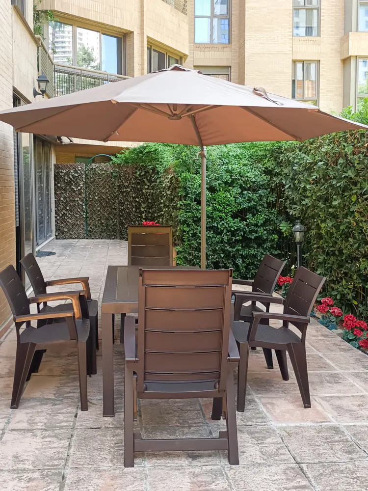 Outdoor patio with brown table and chairs under a large beige umbrella, surrounded by green bushes and red flowers.
