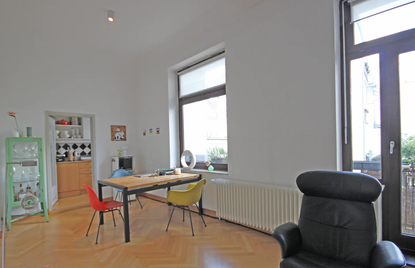 Bright living room with wood floors, white walls, and large windows. A table with colorful chairs sits near a green cabinet and a black leather chair.