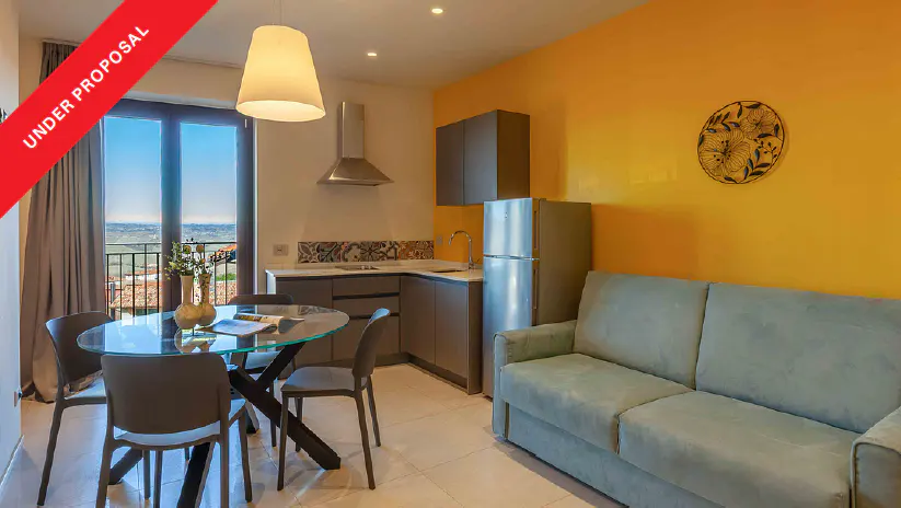 Modern studio apartment with a light blue sofa, glass dining table, kitchenette, and a bright yellow accent wall.