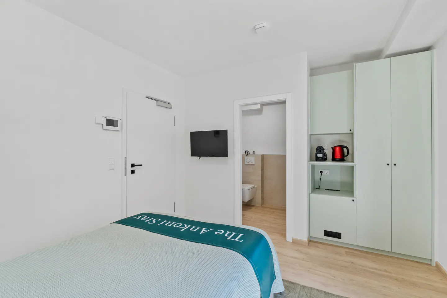 A bright hotel room with a bed, TV, and a glimpse into the bathroom. A light green cabinet with a coffee maker and kettle stands against the wall.