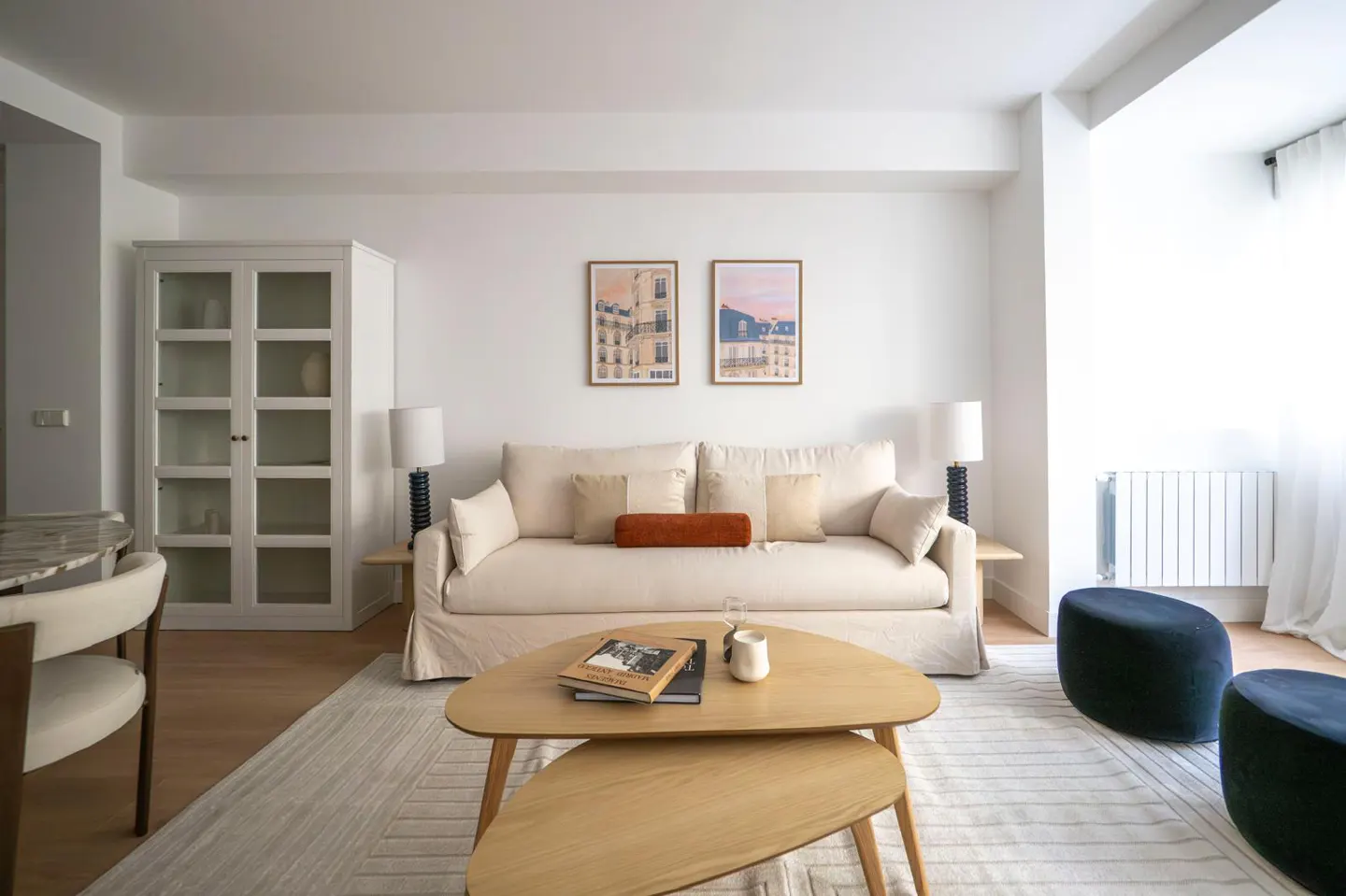 Bright living room with white walls, a beige sofa with pillows, and a light wood coffee table on a patterned rug. Two framed prints hang above the sofa.