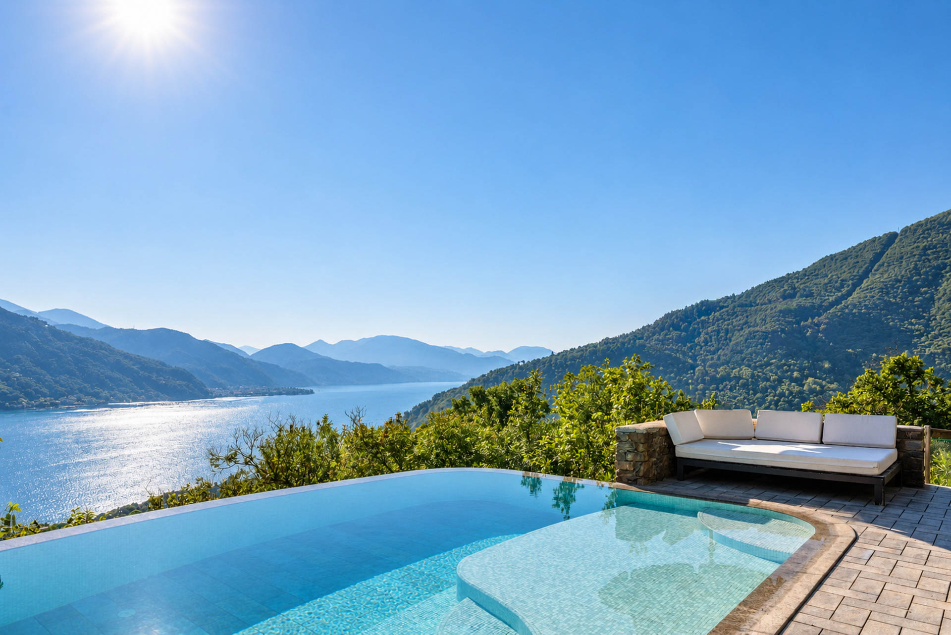 Infinity pool overlooking a sunlit lake and mountains, with a white lounge sofa on a stone patio under a clear blue sky. [AI-generated text]