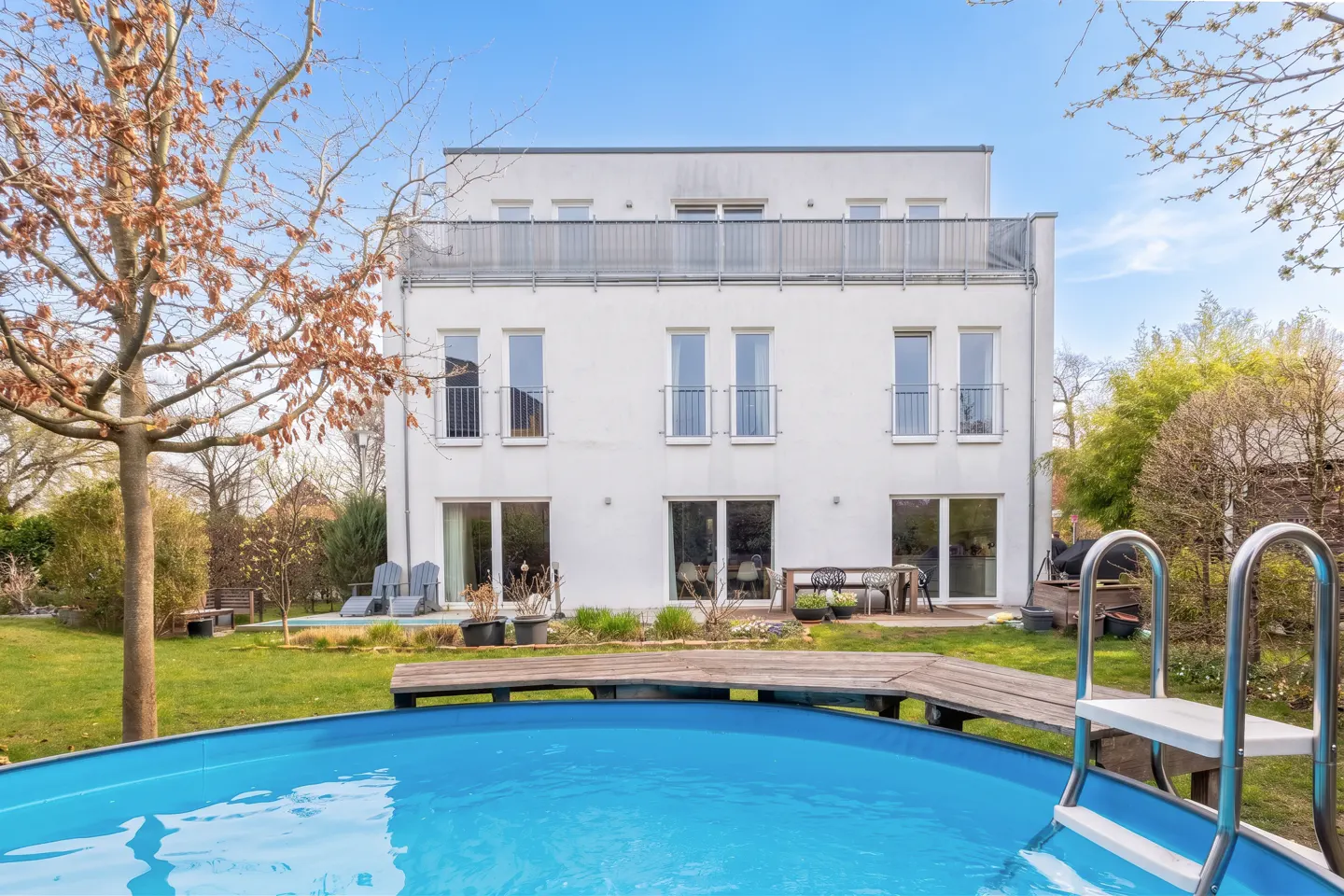 Backyard view of a modern three-story white house with a blue above-ground pool and a wooden deck.