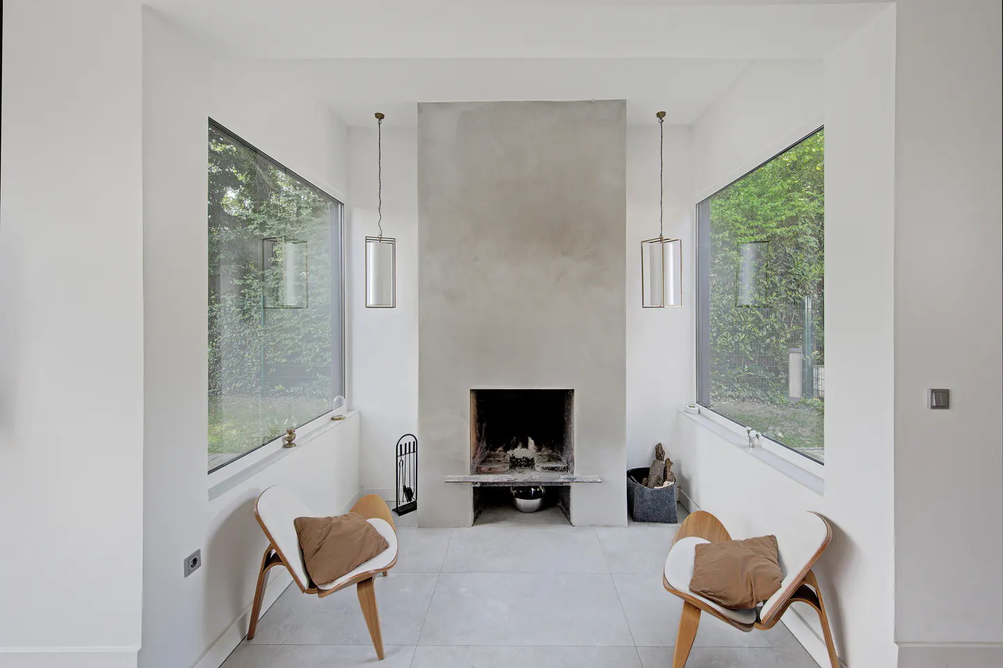 Modern living room with a central concrete fireplace, two white lounge chairs, and large windows overlooking greenery.