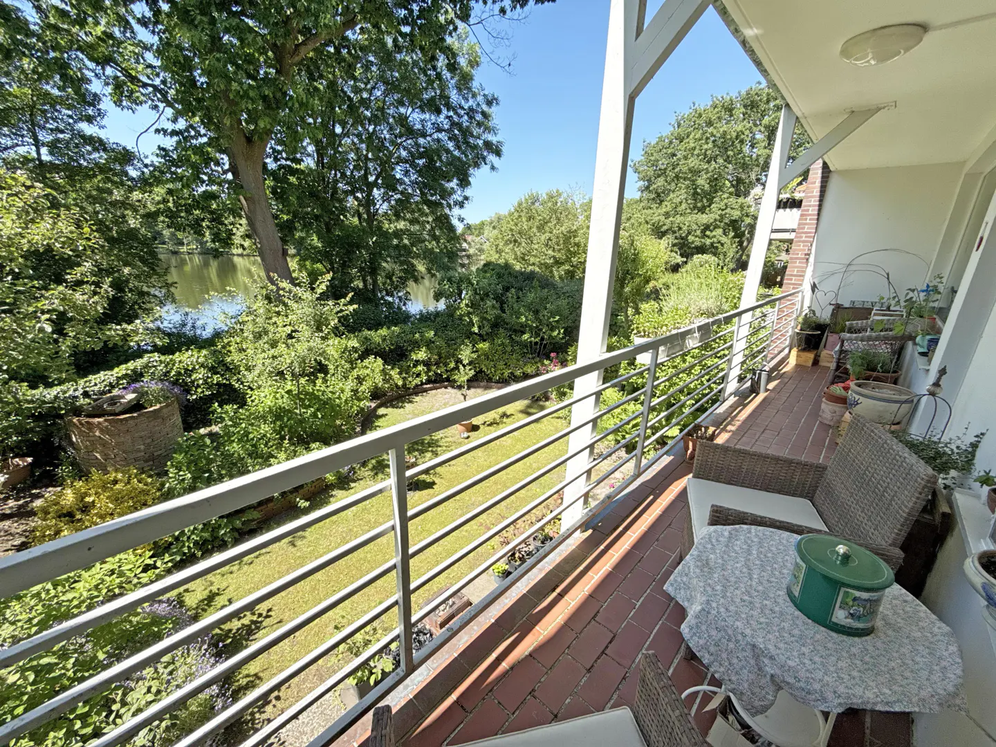 Balcony with wicker chairs and a small table overlooking a lush green garden and a calm lake on a sunny day.