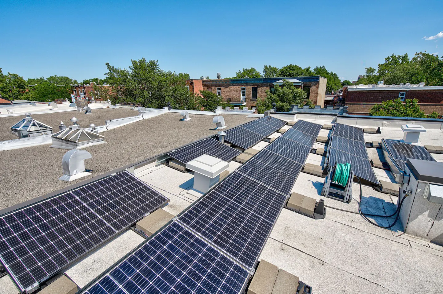 Solar panels on a flat roof with skylights and vents, against a blue sky and trees.