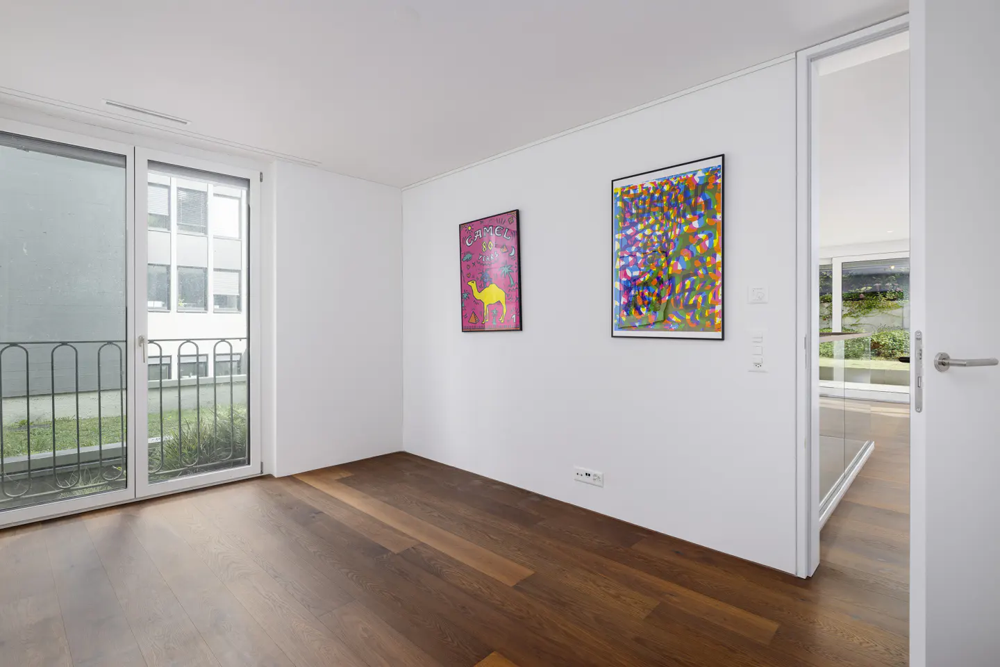 Bright, empty room with hardwood floors, white walls, and two colorful framed posters. A glass door leads to another room with a view of greenery.