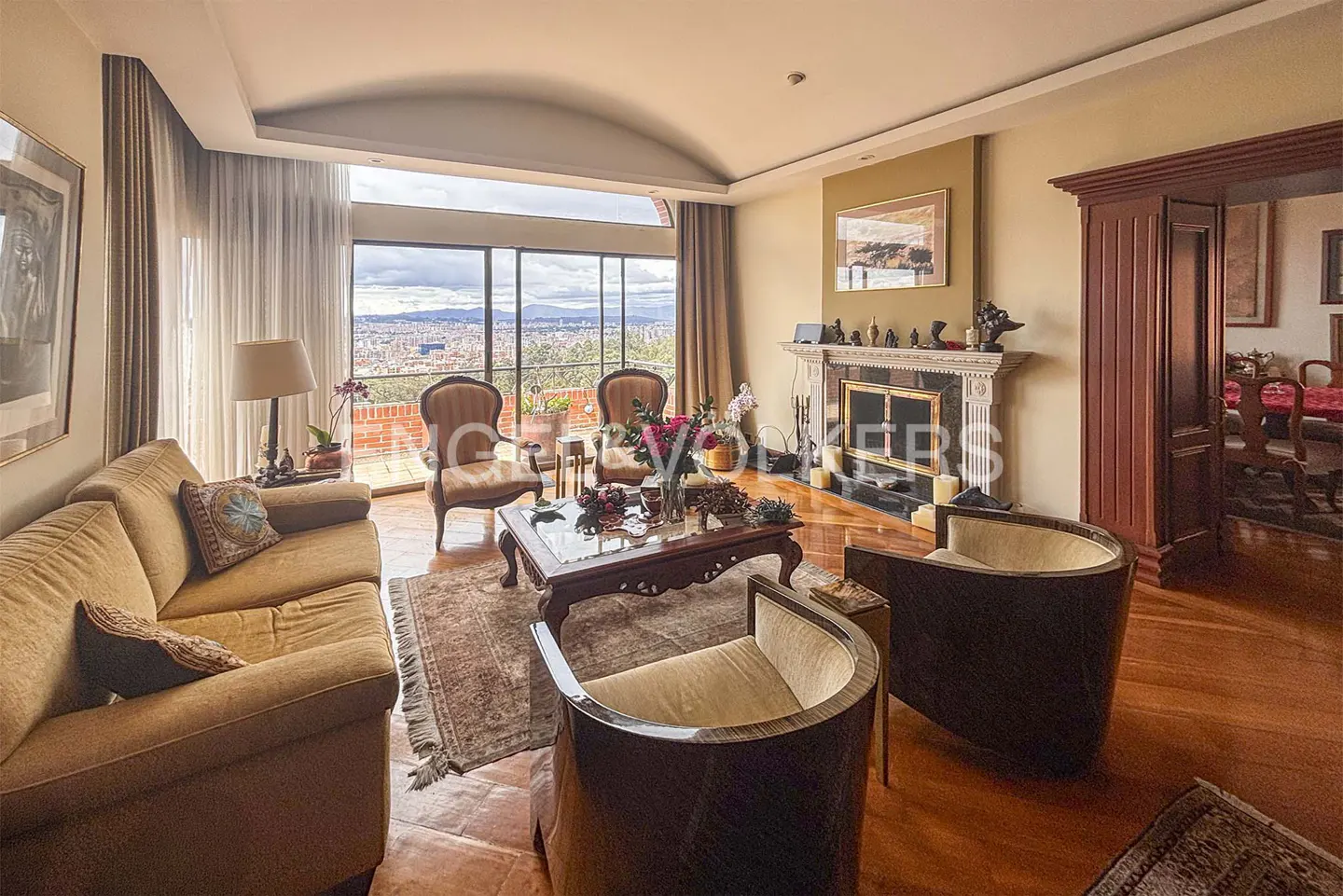 Living room with a tan sofa, dark wood chairs, and a fireplace. A large window shows a city view.