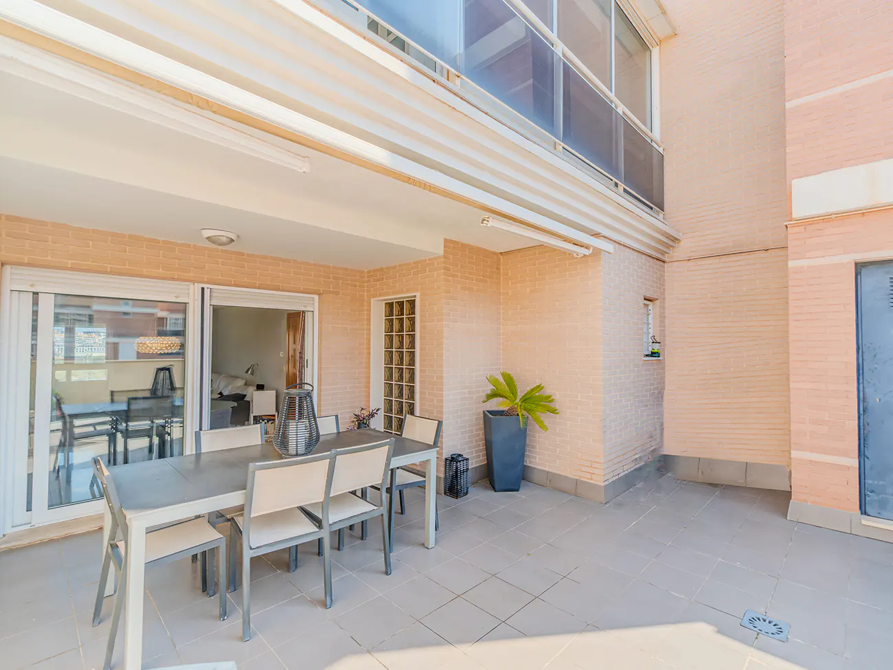 Spacious outdoor patio with a dining table, chairs, and a potted plant against a light brick wall.