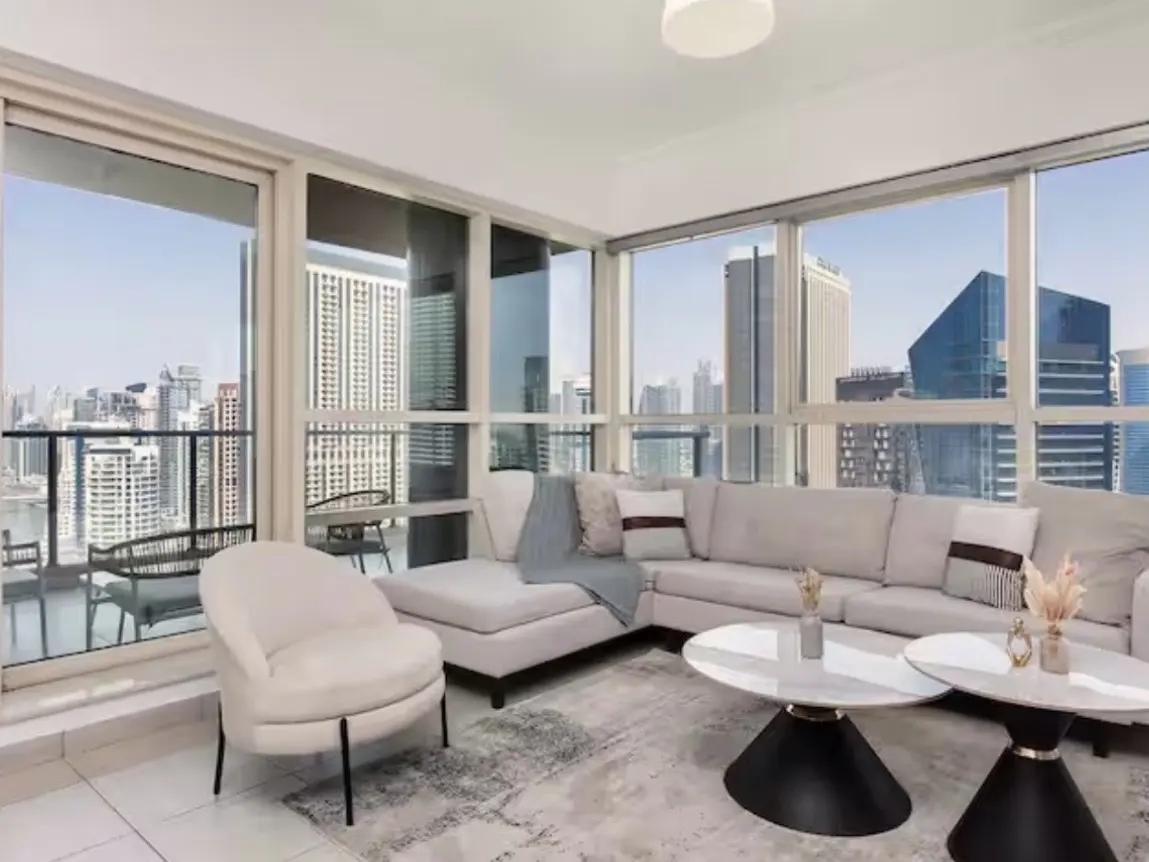 Bright living room with city view. Beige sectional sofa, white chair, and black/white tables on a patterned rug. Large windows show a cityscape.