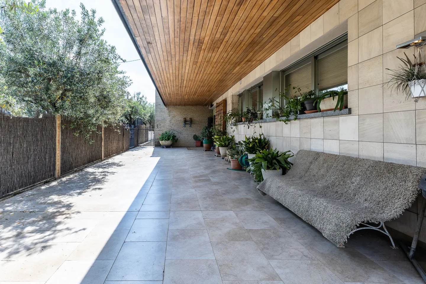 Covered patio with a bench, potted plants, and a wood ceiling. A tree and fence are on the left.