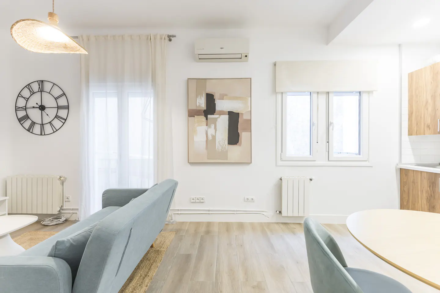 Bright living room with light blue sofa, wood floors, and white walls. A large clock hangs near a window with sheer curtains. An abstract painting is centered above the sofa.