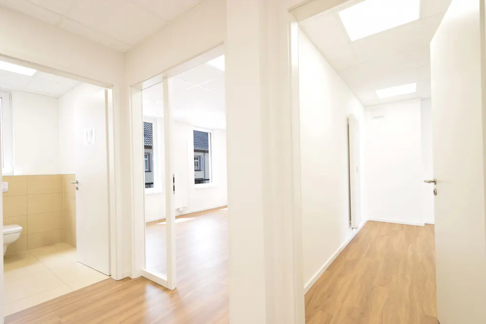Bright, modern office hallway with light wood floors, white walls, and multiple doorways leading to empty rooms.