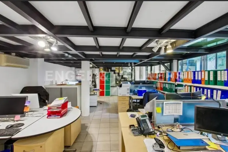 Bright, open office space with desks, computers, and colorful binders on shelves. Black beams support the white ceiling.