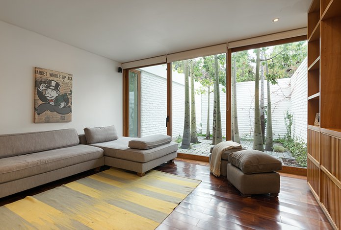 Living room with gray sectional sofa, yellow rug, and a view of trees through a large window. A Monopoly poster hangs on the wall.