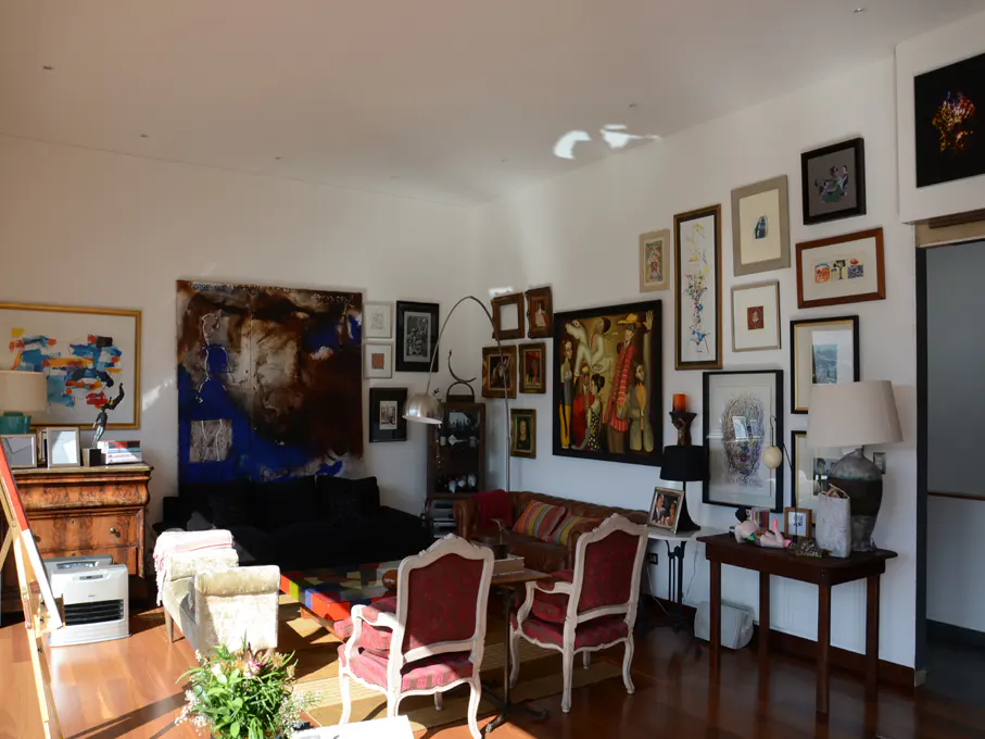 Bright living room with hardwood floors, white walls, and eclectic art collection. Red chairs face a dark sofa. Sunlight streams in.