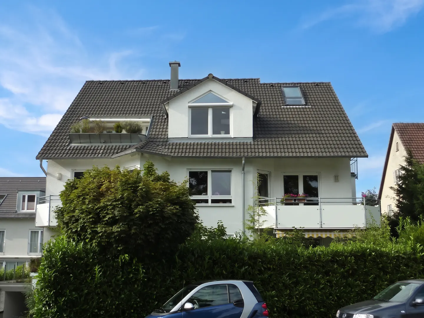 A white two-story house with a gray tiled roof, chimney, and dormer windows, partially obscured by green bushes. A blue car is parked in front.
