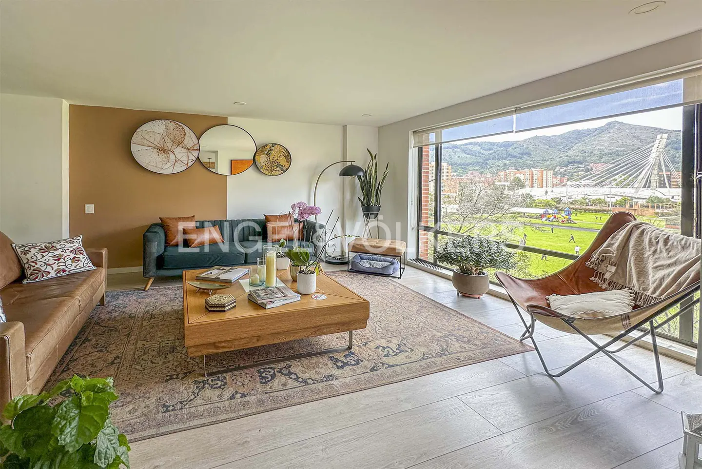 Modern living room with a green sofa, leather armchair, wooden coffee table, and large windows overlooking a city park.
