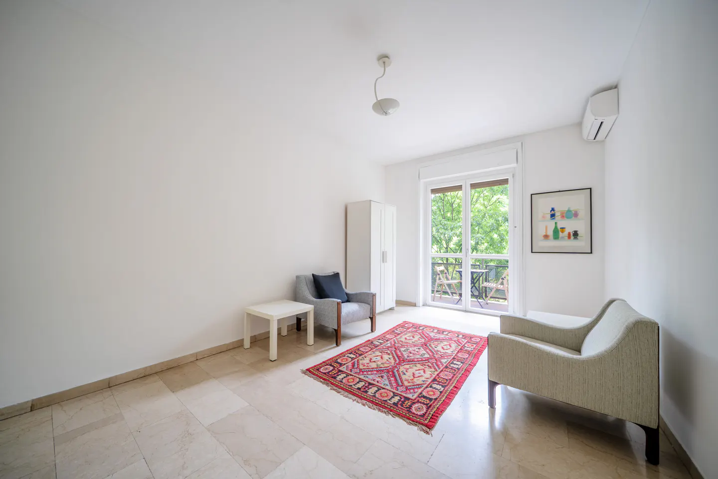 Bright, airy room with white walls, marble floor, and a red patterned rug. Features a gray armchair, chaise lounge, and balcony with green trees visible.